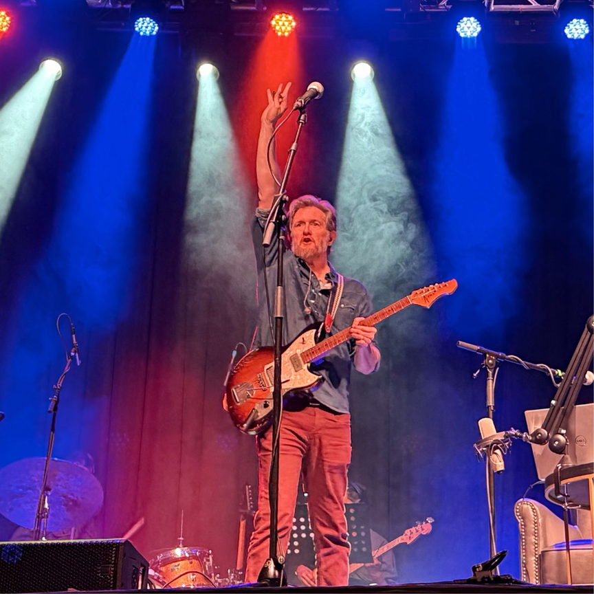 Lindsay Graham playing electric guitar and raising a hand on stage with colourful spotlights and smoke.