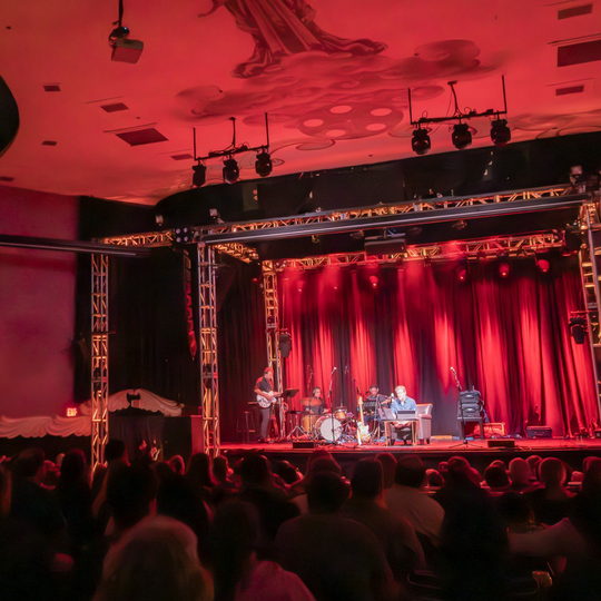 Lindsay Graham and the band performing on stage with red curtains and stage lighting in front of a seated audience.