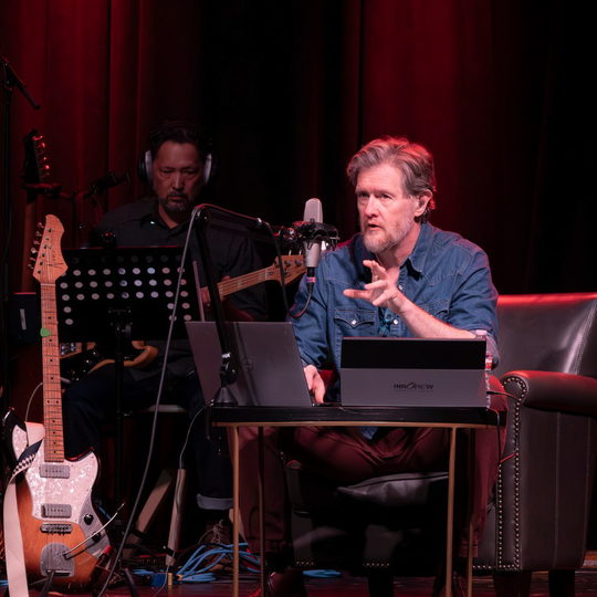 Lindsay Graham speaking into a microphone with a laptop on the table while a band member plays electric guitar.