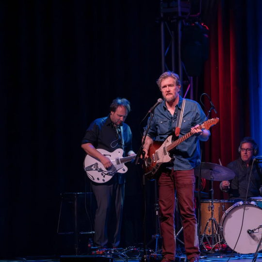 Lindsay Graham and the band on stage performing with electric guitars and drums under blue and red lights.